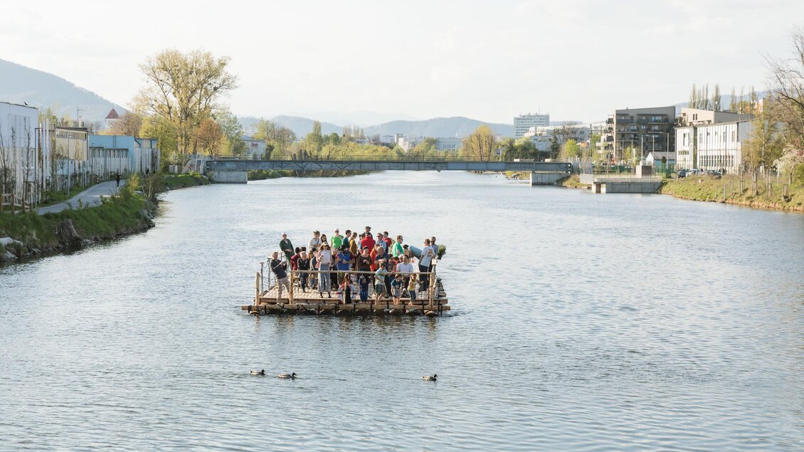 Eine Gruppe von Menschen steht auf einem Floß auf einem ruhigen Fluss. Die Umgebung ist grün und urban, mit Gebäuden im Hintergrund. | © Die Flösserei