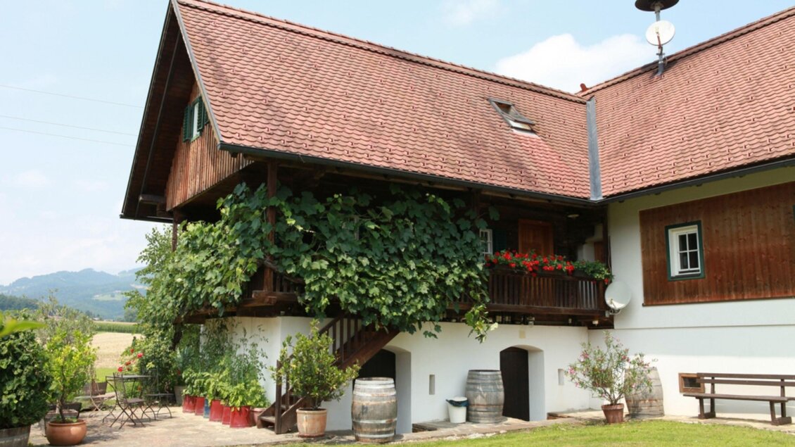 Ein rustikales Haus mit einem roten Dach, umgeben von Grünpflanzen. Im Vordergrund gibt es eine gepflegte Wiese und einige Holzfässer.