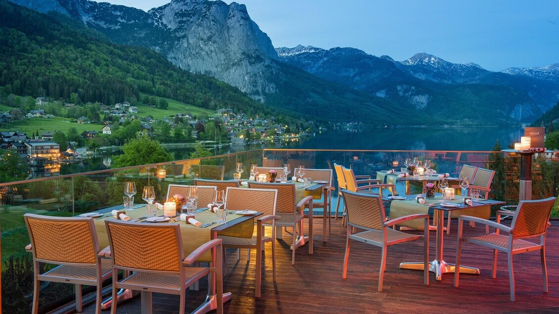 Eine elegante Terrasse mit Tischen und Stühlen bietet einen atemberaubenden Blick auf die Berge und den See. Die sanfte Beleuchtung schafft eine gemütliche Atmosphäre bei Dämmerung. | ©  MONDI Hotels & Resorts