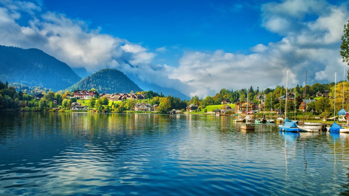 Ein ruhiger See mit sanften Wellen und umliegenden Bergen. Im Vordergrund sind kleine Boote und ein klarer blauer Himmel sichtbar. | ©  MONDI Hotels & Resorts