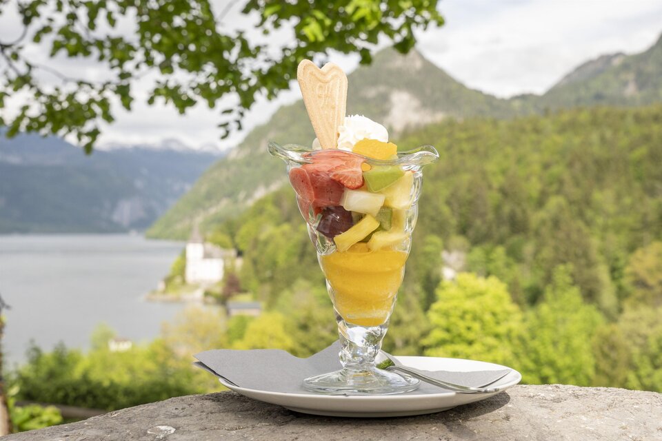 Ein bunter Obstsalat in einem Glas mit Sahne und einer Waffelspitze. Im Hintergrund sind grüne Berge und ein Wassersee zu sehen. | © MONDI Resort