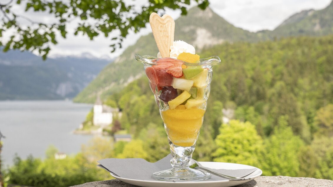 Ein bunter Obstsalat in einem Glas mit Sahne und einer Waffelspitze. Im Hintergrund sind grüne Berge und ein Wassersee zu sehen. | © MONDI Resort