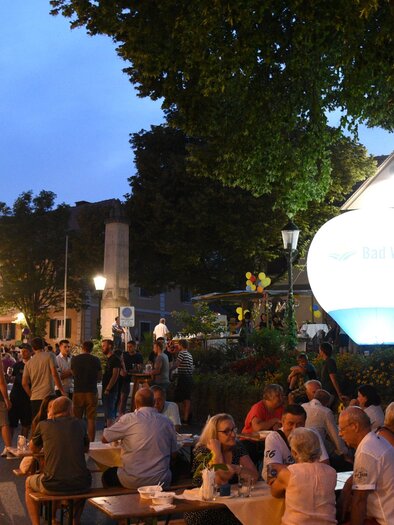 Eine lebhafte Abendveranstaltung mit vielen Menschen, die an Tischen draußen sitzen. Im Hintergrund leuchtet ein großer, runder Ballon und es gibt Bäume und Gebäude in der Umgebung. | © Lederer
