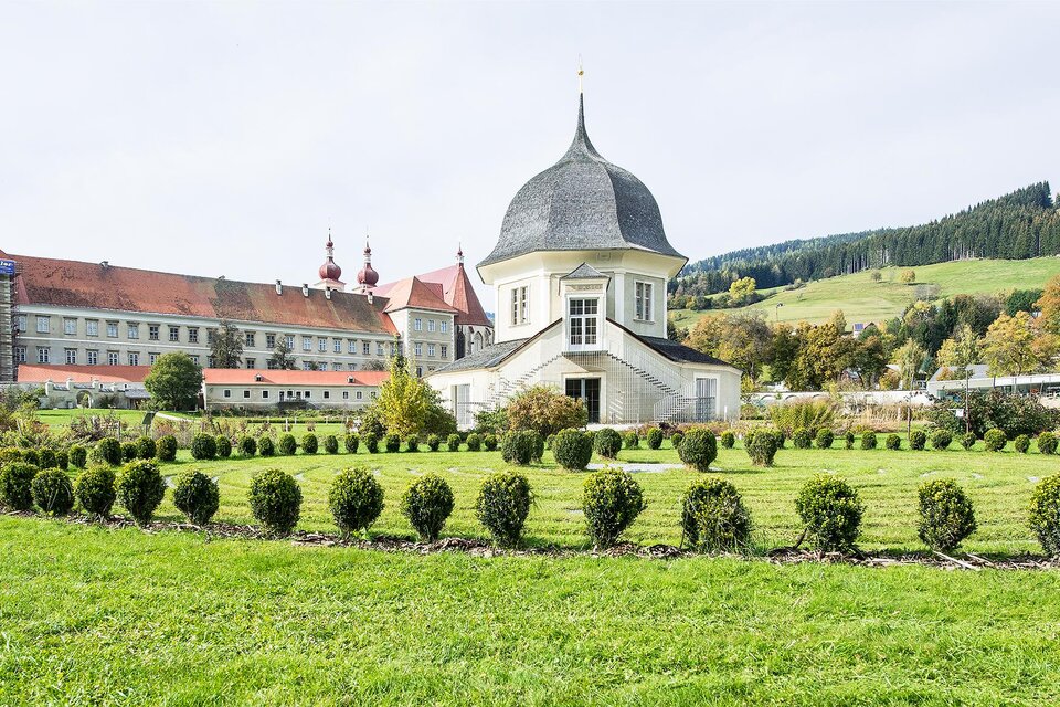 Ein hübsches Gartenareal mit einem weißen Gebäude und einer markanten Kuppel. Im Hintergrund sind sanfte Hügel und historische Gebäude zu sehen. | © Tv Murau