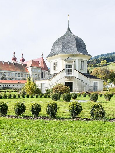 A beautiful garden area featuring a white building with a distinctive dome. In the background, gentle hills and historic buildings can be seen. | © Tv Murau