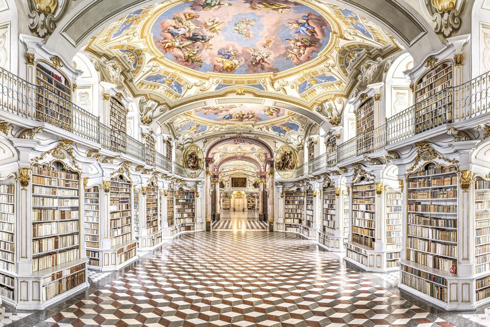 Eine prächtige Bibliothek mit kunstvollen Deckenfresken und regalen voller Bücher. Der Boden ist mit einem auffälligen Schachbrettmuster gestaltet.
