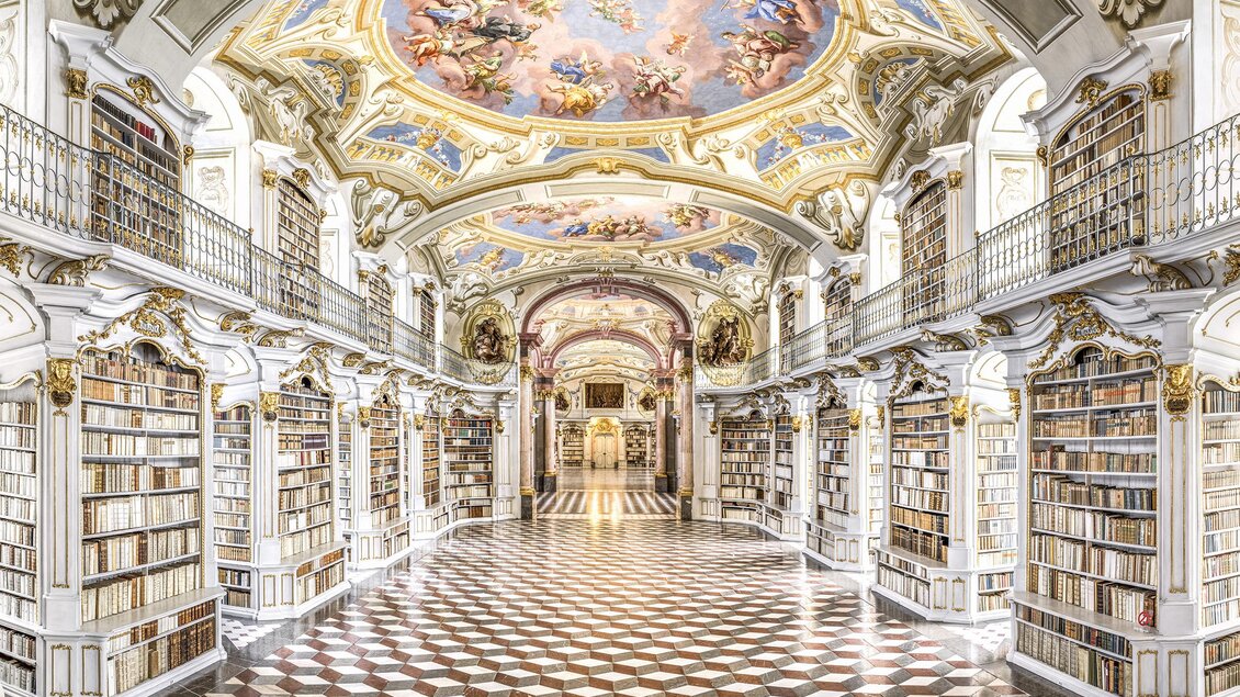 Eine prächtige Bibliothek mit kunstvollen Deckenfresken und regalen voller Bücher. Der Boden ist mit einem auffälligen Schachbrettmuster gestaltet.