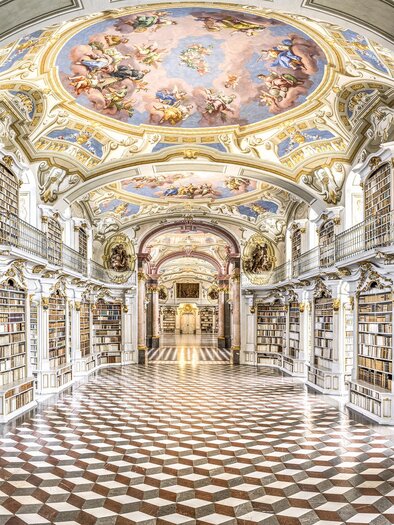 Eine prächtige Bibliothek mit kunstvollen Deckenfresken und regalen voller Bücher. Der Boden ist mit einem auffälligen Schachbrettmuster gestaltet.