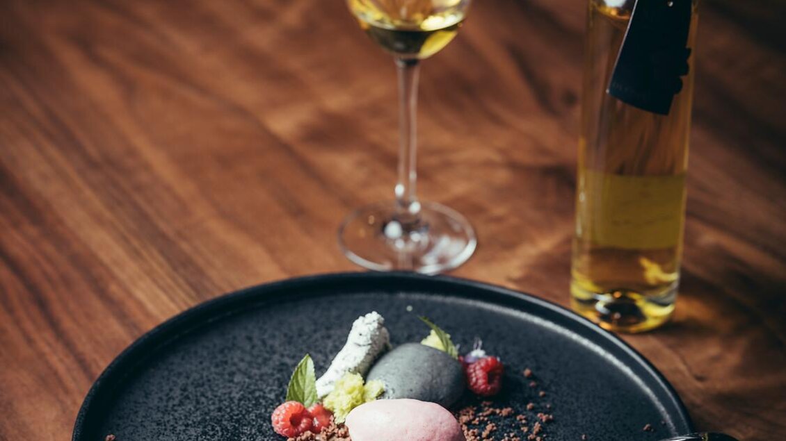 Ein elegantes Dessert auf einem schwarzen Teller, garniert mit verschiedenen Sorbets, frischen Beeren und essbaren Blumen. Daneben steht ein Glas Wein und eine Flasche.