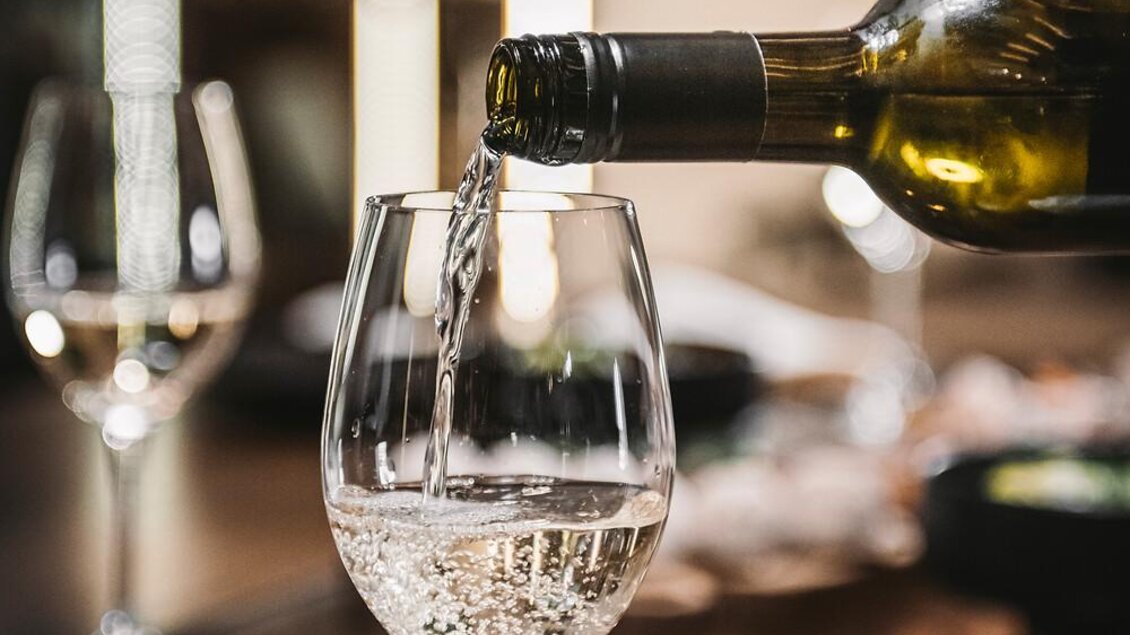 Ein Weinglas wird mit weißem Wein gefüllt. Im Hintergrund sind Kerzen und weitere Gläser sichtbar.