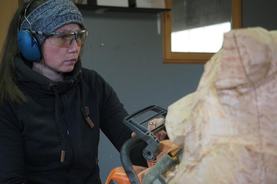Eine Frau arbeitet konzentriert mit einer Kettensäge an einem Holzprojekt. Sie trägt Hörschutz und eine Mütze, während sie einen Teil der Holzskulptur formt.