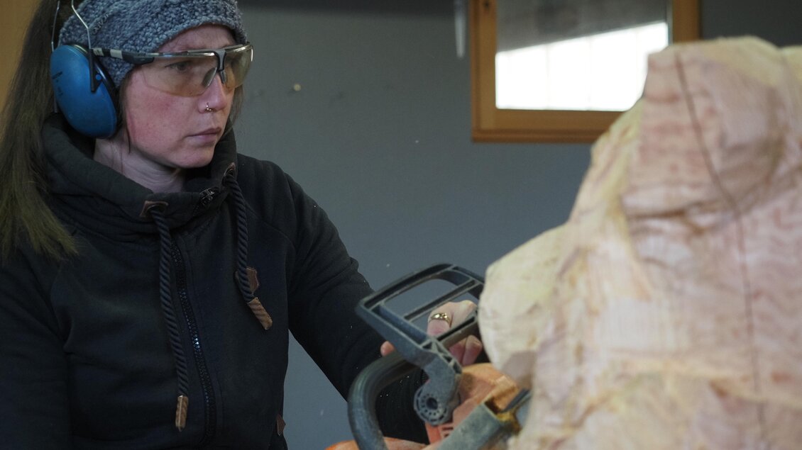 Eine Frau arbeitet konzentriert mit einer Kettensäge an einem Holzprojekt. Sie trägt Hörschutz und eine Mütze, während sie einen Teil der Holzskulptur formt.