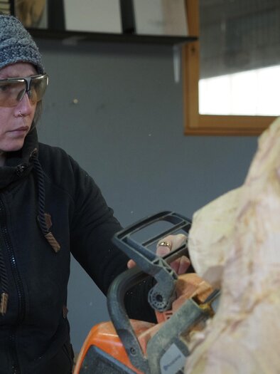 Eine Frau arbeitet konzentriert mit einer Kettensäge an einem Holzprojekt. Sie trägt Hörschutz und eine Mütze, während sie einen Teil der Holzskulptur formt.