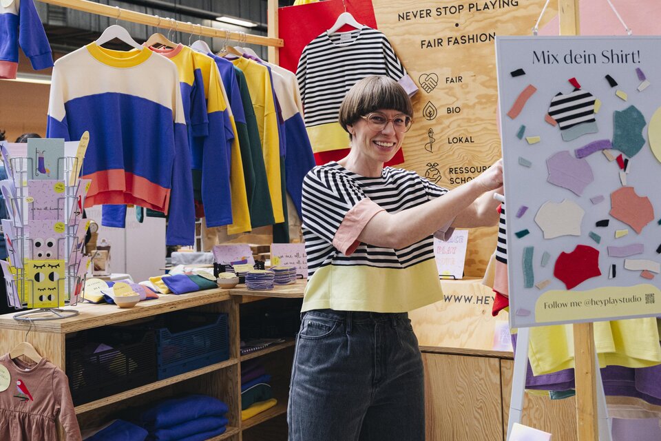 Eine Frau gestaltet an einem Stand ein Shirt, umgeben von farbenfroher Kleidung beim Kunst- und Designmarkt in Graz. | © Franziska Freiwald