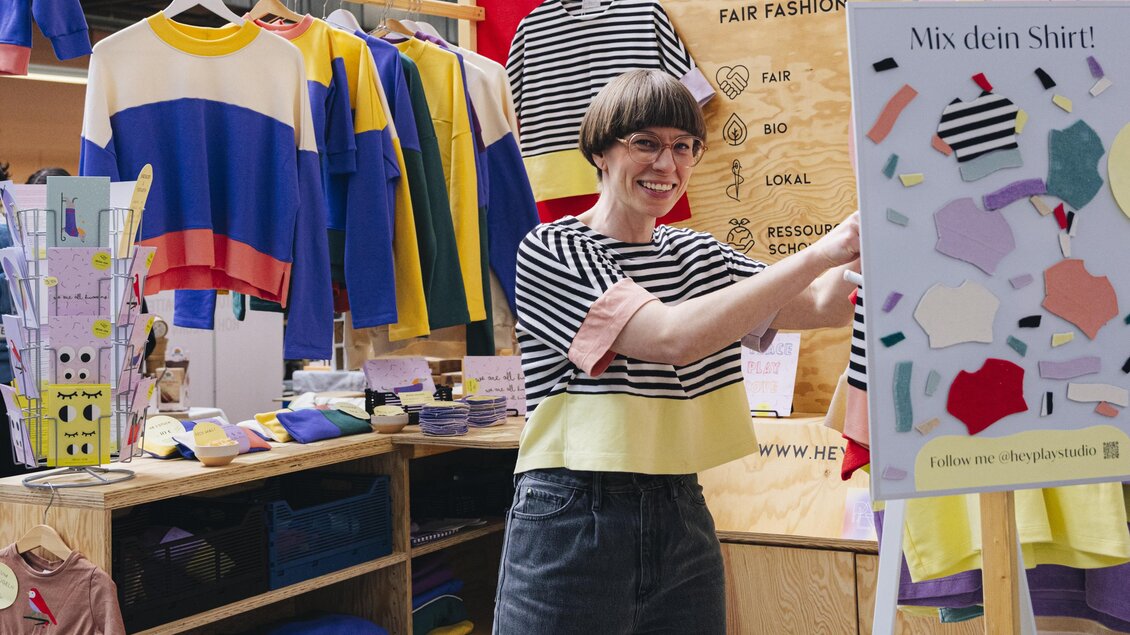Eine Frau gestaltet an einem Stand ein Shirt, umgeben von farbenfroher Kleidung beim Kunst- und Designmarkt in Graz. | © Franziska Freiwald