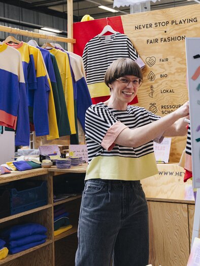 Eine Frau gestaltet an einem Stand ein Shirt, umgeben von farbenfroher Kleidung beim Kunst- und Designmarkt in Graz. | © Franziska Freiwald