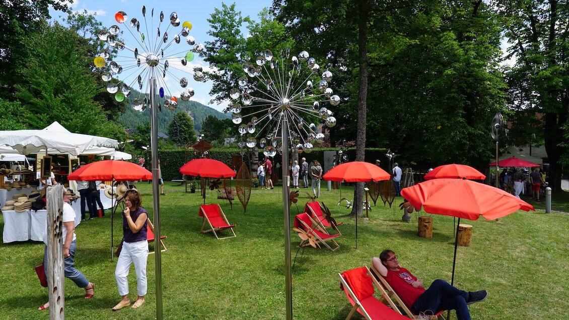 Ein schöner Garten mit bunten Windspielen und roten Sonnenschirmen. Besucher entspannen auf Liegestühlen und genießen die Atmosphäre. | © Andrea Lackner