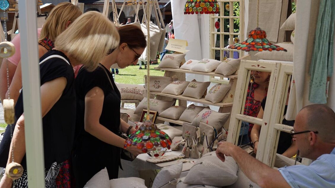 Zwei Frauen betrachten handgefertigte Produkte auf einem Marktstand. Im Hintergrund sind weitere Besucher und dekorative Artikel sichtbar. | © Andrea Lackner