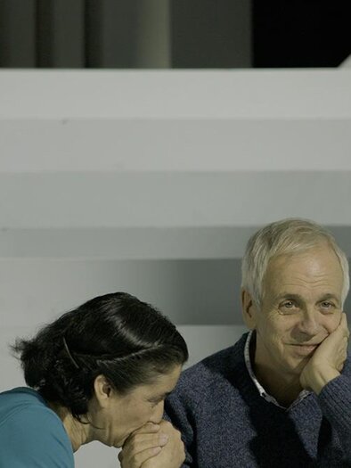An older man is sitting thoughtfully on a staircase while a woman is whispering confidentially into his ear. The scene is simple and focused on the two individuals. | ©  Polyfilm