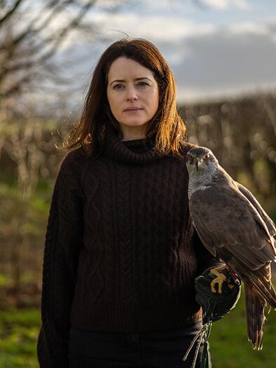 A woman stands outside and holds a falcon on her wrist. In the background, there are trees and a slightly cloudy sky. | © Panda Film
