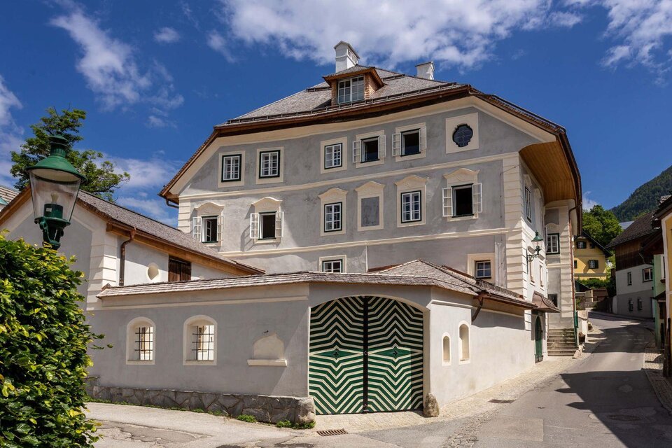 Das historische Podenhaus in Bad Aussee unter strahlend blauem Himmel. Es ist ein schönes großes Stadthaus mit vielen Fenstern, die Fassadenfarbe ist überwiegend weiß mit einigen historischen Elementen. Im Vordergrund ist das markante grünweiße Eingangstor zum Innenhof zu sehen.< | © Christian Cavalcante dos Santos