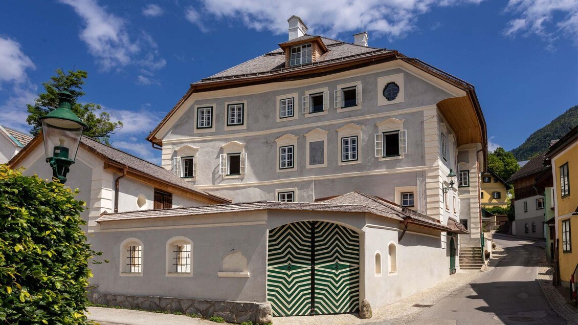 Das historische Podenhaus in Bad Aussee unter strahlend blauem Himmel. Es ist ein schönes großes Stadthaus mit vielen Fenstern, die Fassadenfarbe ist überwiegend weiß mit einigen historischen Elementen. Im Vordergrund ist das markante grünweiße Eingangstor zum Innenhof zu sehen.< | © Christian Cavalcante dos Santos