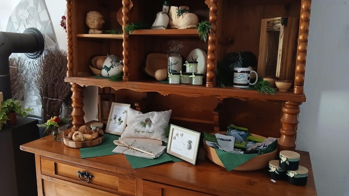 Ein Holzschrank mit verschiedenen dekorativen Gegenständen und Geschenken. Oben sind Keramiken und Gläser zu sehen, während unten einige Kissen und grünes Dekor liegen. | © Michelle Friedam