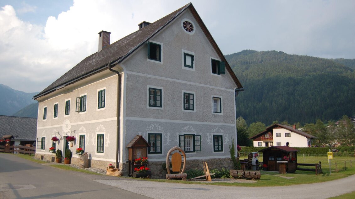 Ein traditionshaus mit einer grauen Fassade und großen Fenstern. Im Hintergrund sind grüne Berge und eine ruhige Landschaft zu sehen. | © Creativ Verein Tragöß