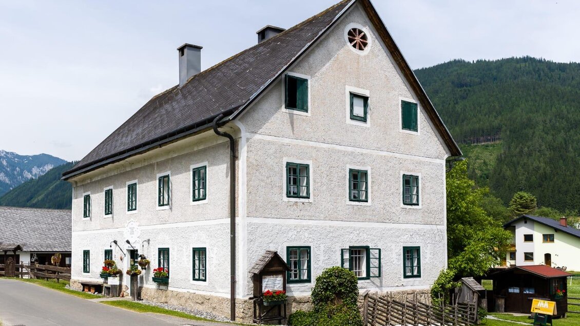 Ein traditionelles, zweigeschossiges Gebäude mit weißen Wänden und grünen Fenstern. Es steht an einer ruhigen Straße in einer malerischen Berglandschaft. | © Creativ Verein Tragöß