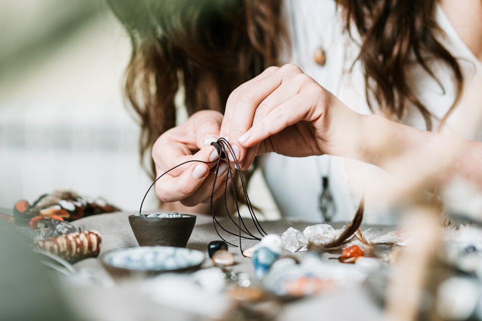 Eine Person fädelt sorgfältig eine Schnur durch einen Draht. Auf dem Tisch liegen verschiedene Schmuckstücke und Materialien. | © adobe stock foto