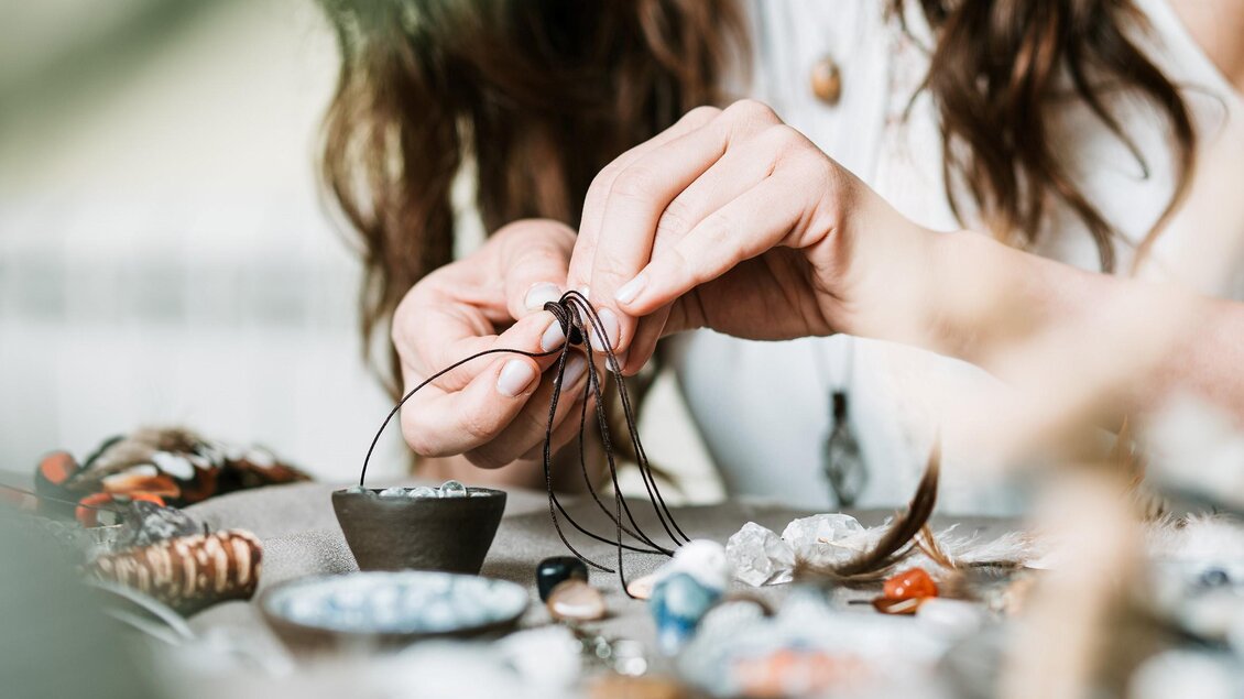 Eine Person fädelt sorgfältig eine Schnur durch einen Draht. Auf dem Tisch liegen verschiedene Schmuckstücke und Materialien. | © adobe stock foto