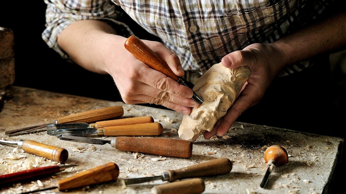 Ein Handwerker schnitzt mit einem Holzwerkzeug an einer Holzfigur. Werkzeuge liegen auf einem Tisch, umgeben von Holzspänen. | © adobe stock foto