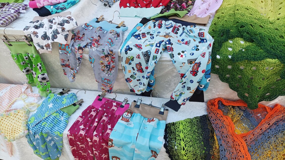 Bunte Kinderhosen und handgehäkelte Tücher liegen auf einem Tisch. Die Kleidung zeigt verschiedene Muster und Farben. | © Stadtgemeinde Hartberg
