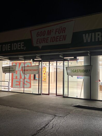 Ein Geschäft mit großen Fenstern und einem transparenten Eingangsbereich. Es ist nachts und die Innenbeleuchtung ist eingeschaltet. | © MitteninZeltweg