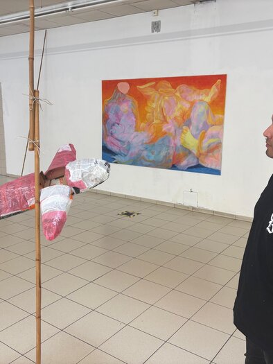 Eine Person steht in einem Raum und schaut auf ein hängendes, rosa Fischkunstwerk. Im Hintergrund ist ein buntes Gemälde zu sehen. | © manifestderleere