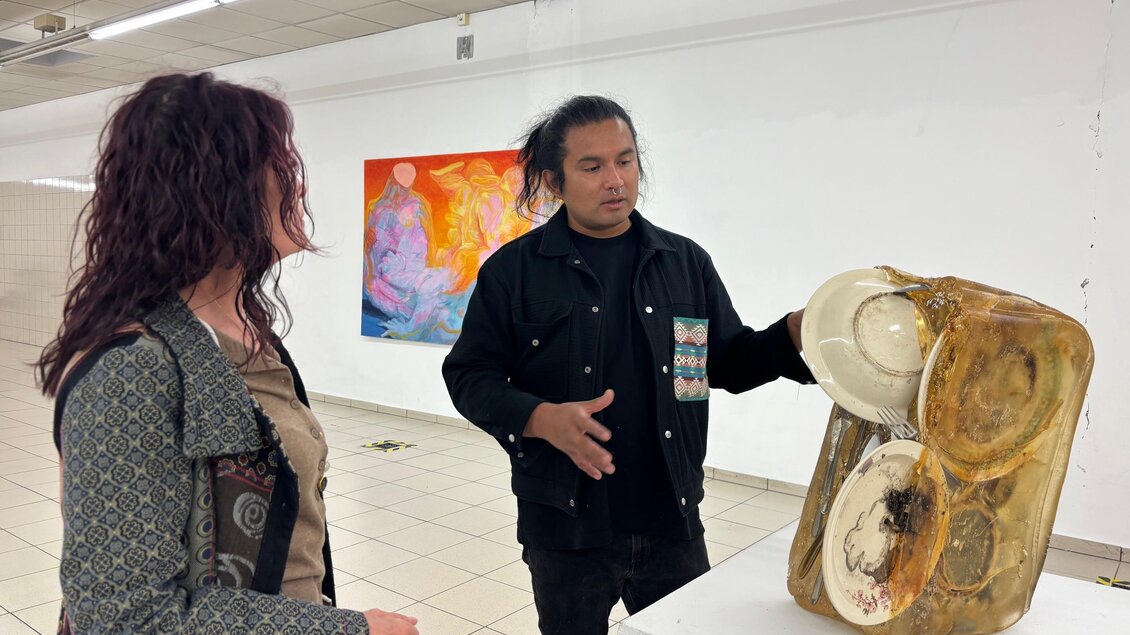 Eine Person erklärt eine Skulptur aus verschiedenen Materialien in einer Galerie. Im Hintergrund sind abstrakte Gemälde zu sehen. | © manifestderleere