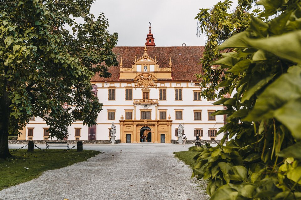 Ein beeindruckendes historisches Gebäude mit einem großen Eingangsbereich und kunstvollen Verzierungen. Umgeben von Bäumen und einem Kiesweg, vermittelt es eine ruhige Atmosphäre. | © _Region Graz_Reisehappen