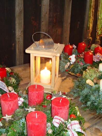 A festive arrangement of candles and lanterns on a wooden table. The red and white candles are surrounded by green wreaths, creating a cozy atmosphere. | © ÖFM