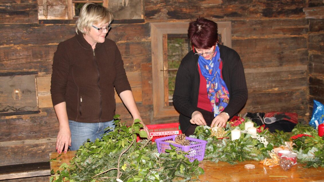 Zwei Frauen arbeiten an einem Tisch mit grünen Pflanzen und Blumen. Im Hintergrund ist eine rustikale Holzwand zu sehen. | © Freilichtmuseum Stübing