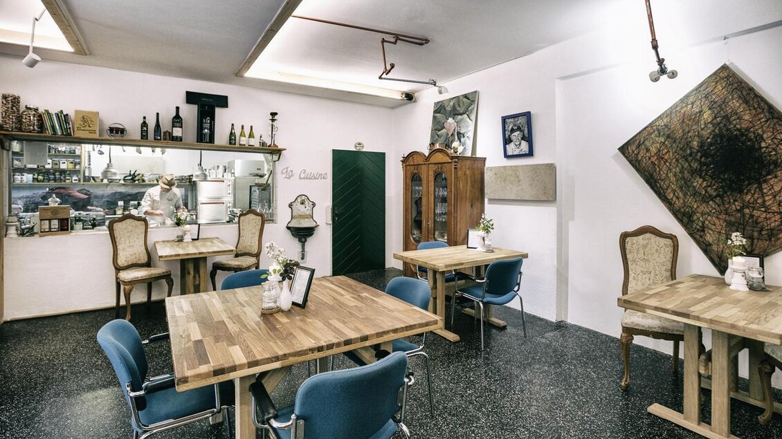 Ein einladendes Restaurant mit Holztischen und blauen Stühlen. Im Hintergrund sind Regale mit Flaschen und eine offene Küchenzeile zu sehen. | © Löke