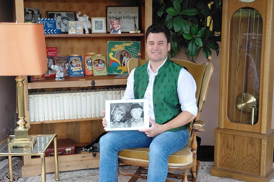 Das Foto zeigt **Anton Nöst-Löwinger**, den Enkel der bekannten österreichischen Volksschauspielerin **Sissy Löwinger**, in einem liebevoll nostalgisch eingerichteten Raum. In seinen Händen hält er eine historische Schwarz-Weiß-Aufnahme, während ihn zahlreiche Fotografien, Bücher, Tonträger und Erinnerungsstücke umgeben. Die große Standuhr, das alte Radio und die klassische Einrichtung verleihen der Aufnahme einen besonderen Charme. Das Bild verbindet auf stimmungsvolle Weise **Familiengeschichte, Theatertradition und die Erinnerung an Sissy Löwinger**. | © Anton Nöst-Löwinger