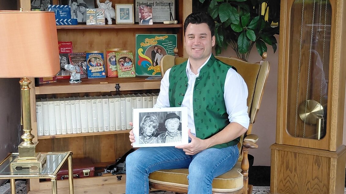 Das Foto zeigt **Anton Nöst-Löwinger**, den Enkel der bekannten österreichischen Volksschauspielerin **Sissy Löwinger**, in einem liebevoll nostalgisch eingerichteten Raum. In seinen Händen hält er eine historische Schwarz-Weiß-Aufnahme, während ihn zahlreiche Fotografien, Bücher, Tonträger und Erinnerungsstücke umgeben. Die große Standuhr, das alte Radio und die klassische Einrichtung verleihen der Aufnahme einen besonderen Charme. Das Bild verbindet auf stimmungsvolle Weise **Familiengeschichte, Theatertradition und die Erinnerung an Sissy Löwinger**. | © Anton Nöst-Löwinger