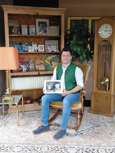 Das Foto zeigt **Anton Nöst-Löwinger**, den Enkel der bekannten österreichischen Volksschauspielerin **Sissy Löwinger**, in einem liebevoll nostalgisch eingerichteten Raum. In seinen Händen hält er eine historische Schwarz-Weiß-Aufnahme, während ihn zahlreiche Fotografien, Bücher, Tonträger und Erinnerungsstücke umgeben. Die große Standuhr, das alte Radio und die klassische Einrichtung verleihen der Aufnahme einen besonderen Charme. Das Bild verbindet auf stimmungsvolle Weise **Familiengeschichte, Theatertradition und die Erinnerung an Sissy Löwinger**. | © Anton Nöst-Löwinger