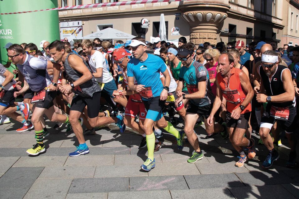 Eine Gruppe von Läufern startet gerade ein Rennen auf einer belebten Straße. Die Teilnehmer tragen vielfältige Sportkleidung und sind voller Energie. | © Pressberger
