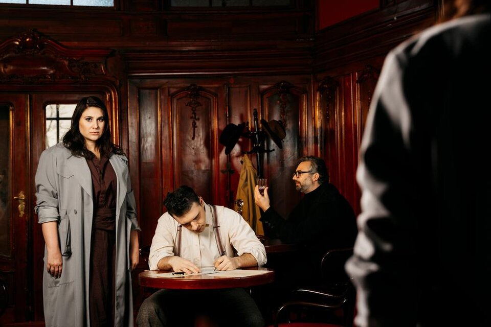 Eine dramatische Szene in einem eleganten Raum. Eine Frau steht im Vordergrund, während zwei Männer am Tisch sitzen, einer von ihnen schreibt. | © Ingo Pertramer_Oper Graz