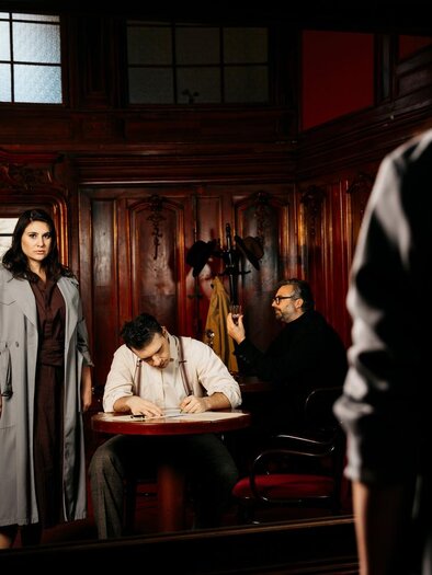 Eine dramatische Szene in einem eleganten Raum. Eine Frau steht im Vordergrund, während zwei Männer am Tisch sitzen, einer von ihnen schreibt. | © Ingo Pertramer_Oper Graz