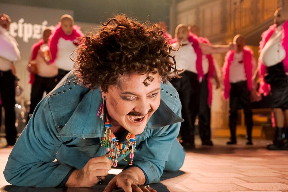 Ein fröhlicher Performer in einem bunten Kostüm führt eine lebhafte Darbietung auf. Im Hintergrund tanzen mehrere Personen in pinken Schals. | © Oper Graz