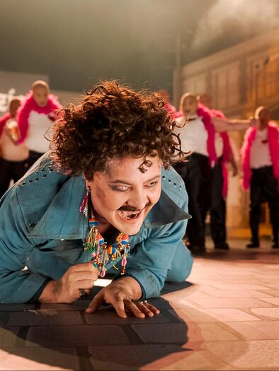 Ein fröhlicher Performer in einem bunten Kostüm führt eine lebhafte Darbietung auf. Im Hintergrund tanzen mehrere Personen in pinken Schals. | © Oper Graz