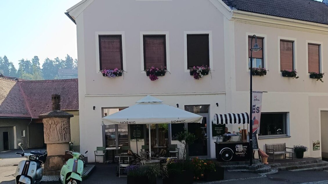 Ein charmantes Gebäude an einer Straße, umgeben von Bäumen. Vor dem Lokal stehen ein paar Fahrzeuge und ein Sonnenschirm. | © Jürgen Atschko