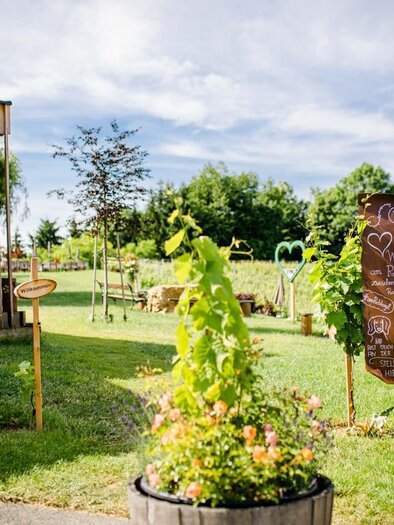Ein malerischer Garten mit bunten Blumen und einem Holzgebäude. Ein Schild zeigt verschiedene Angebote an, umgeben von üppigem Grün. | © Buschenschank Tinnauer